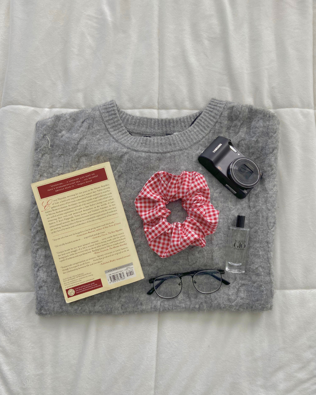 Gray sweater with a book, red scrunchie, camera, and glasses on a white surface