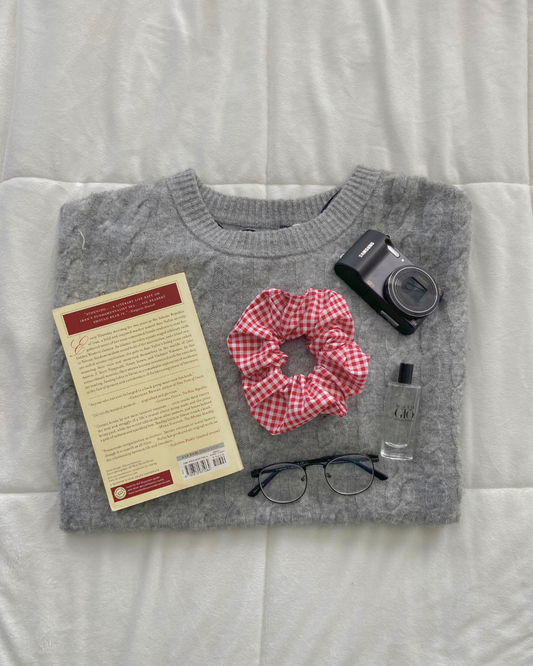 Gray sweater with a book, red scrunchie, camera, and glasses on a white surface
